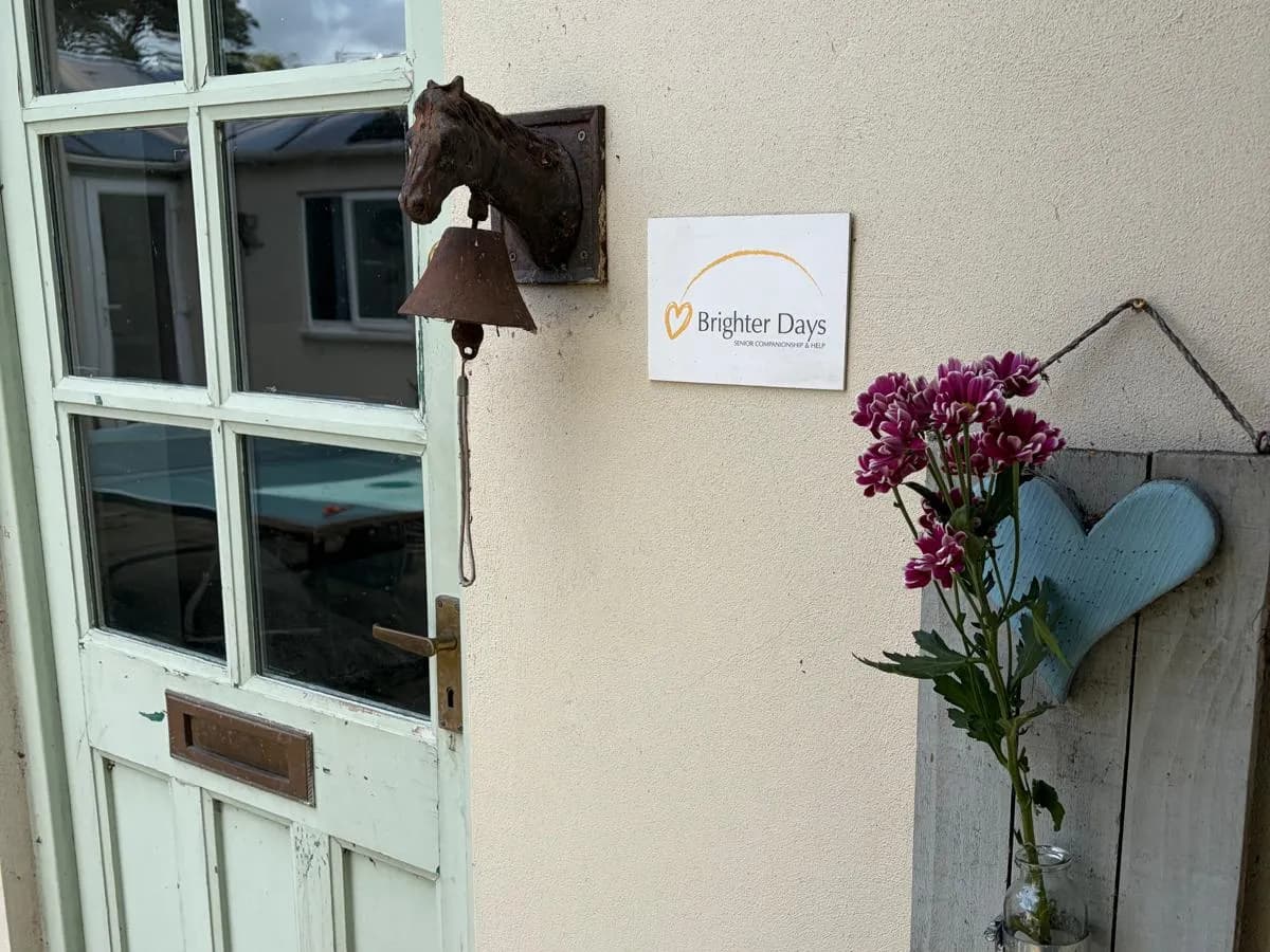 The front door of Brighter Days Companions: a sage green cottage door with a cast-iron horse bell beside it, the Brighter Days sign on the wall, and a wooden heart holding a jar of fresh pink flowers