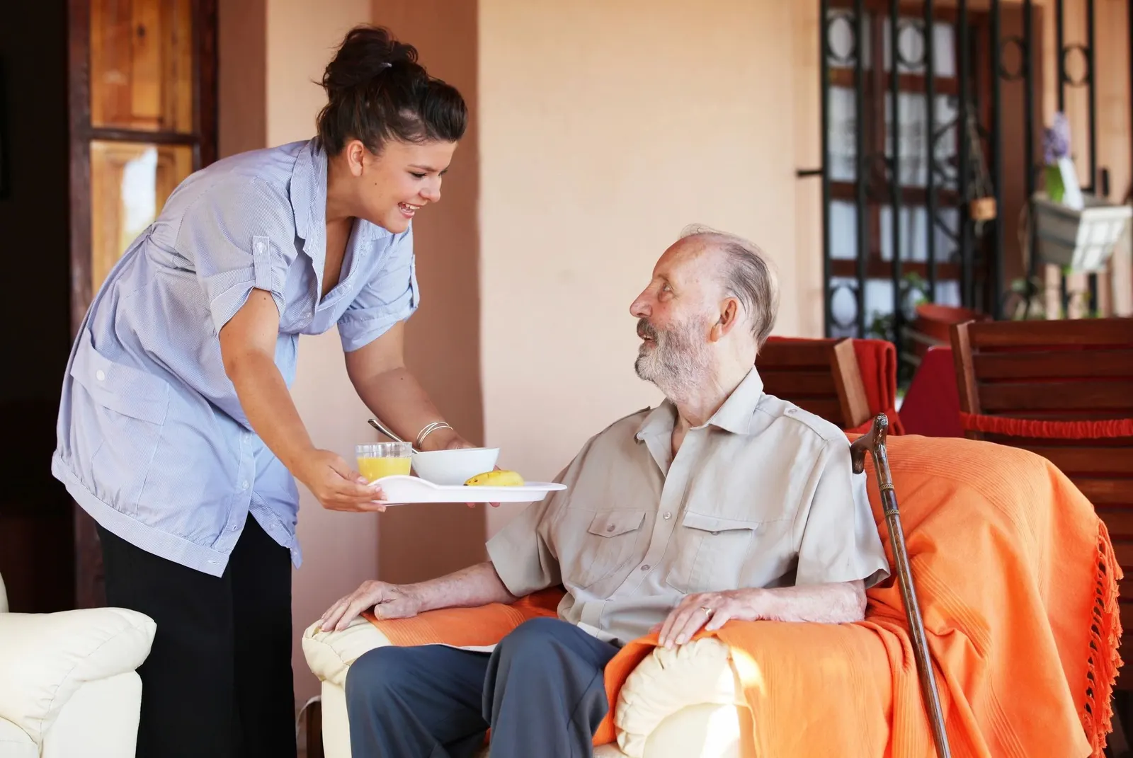 A carer bringing a tray of food to an older man sitting in an armchair