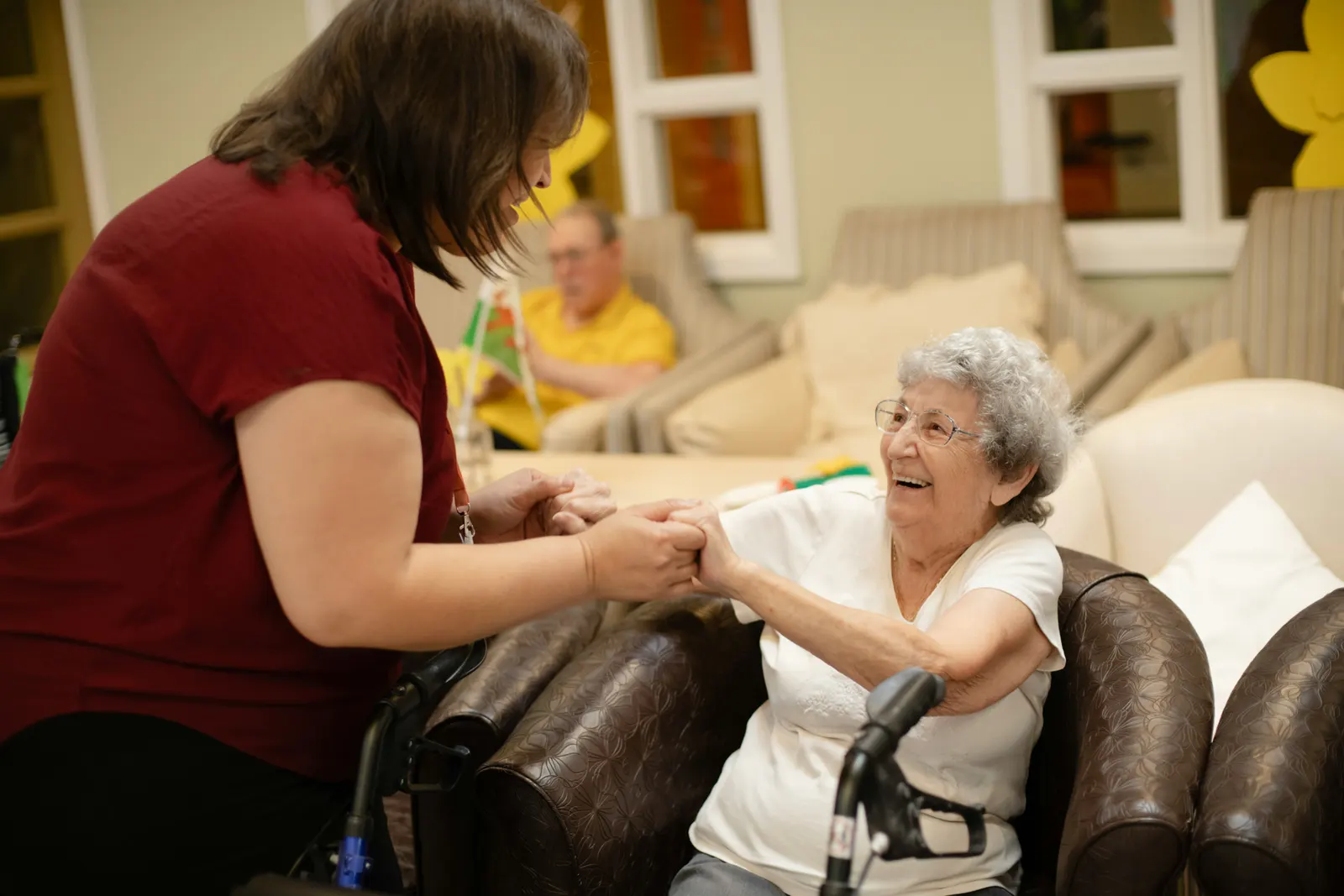 A carer holding both hands of a smiling older woman sitting in an armchair at home